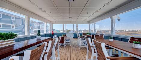 A covered outdoor dining area with wooden tables and white chairs, large windows offering views of the beach and buildings.
