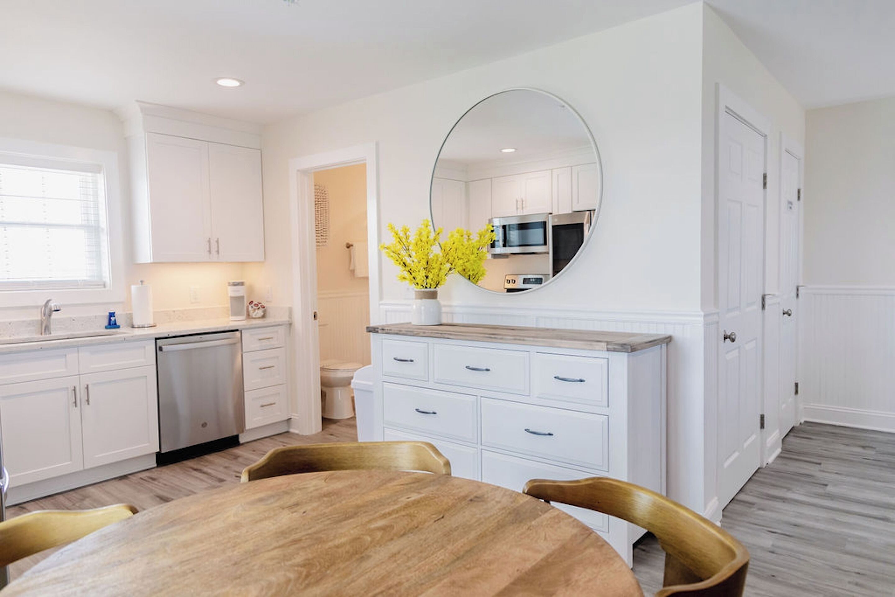A modern kitchen with white cabinetry, a wooden dining table, and a large mirror above the countertop.