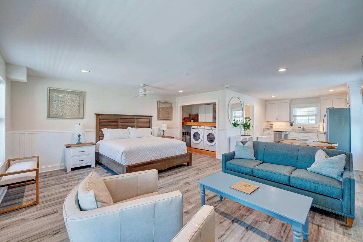Ein modernes Hotelzimmer mit Bett, Sofa und kleinem Esstisch. Im Hintergrund sind eine Waschmaschine und eine Küchenzeile erkennbar.