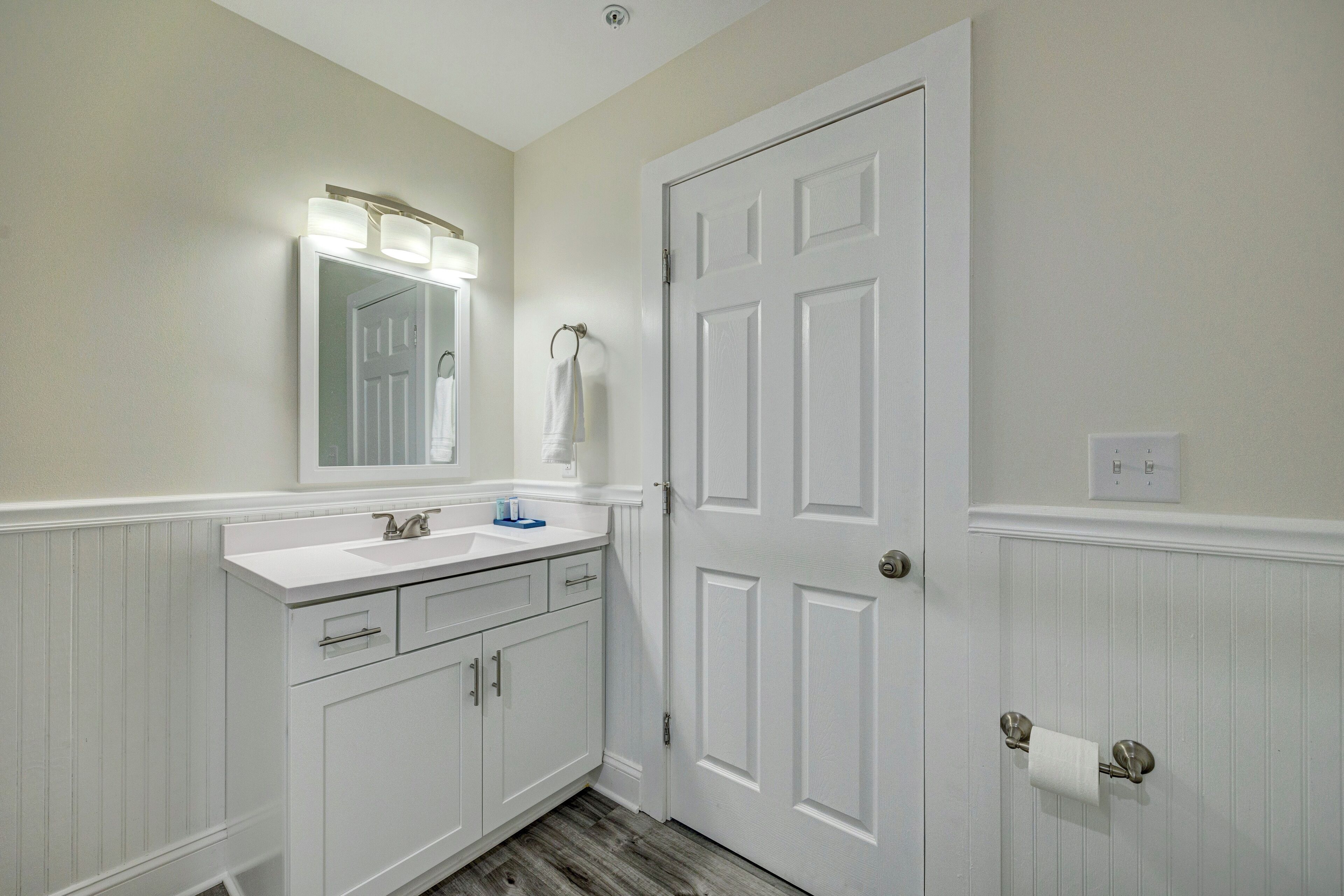A bathroom with a white vanity, a mirror with lights, a door, and a towel rack.