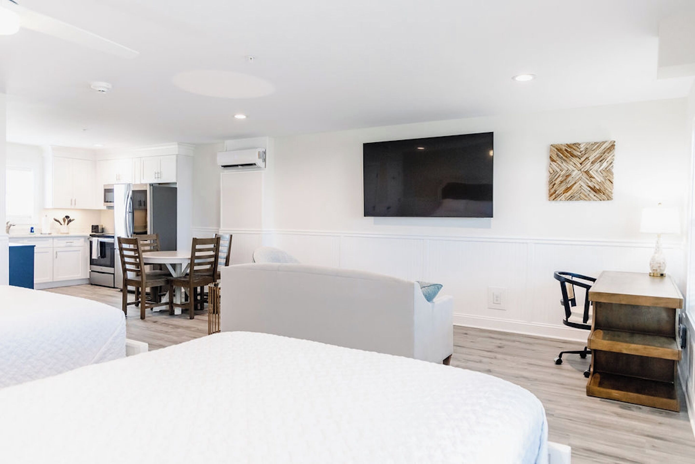 A modern hotel room with a kitchenette, a large flat-screen TV, a comfortable sofa, and a desk with a lamp.