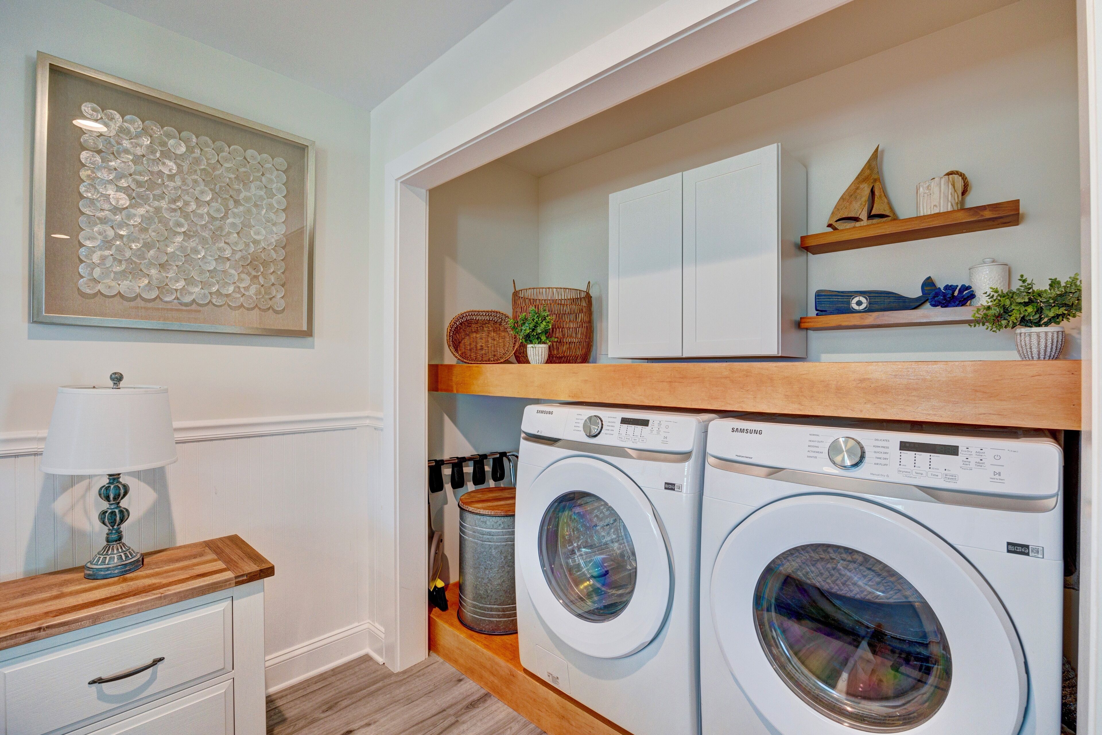 A laundry room with two white washing machines, a wooden shelf with decorative items, and a framed artwork on the wall.