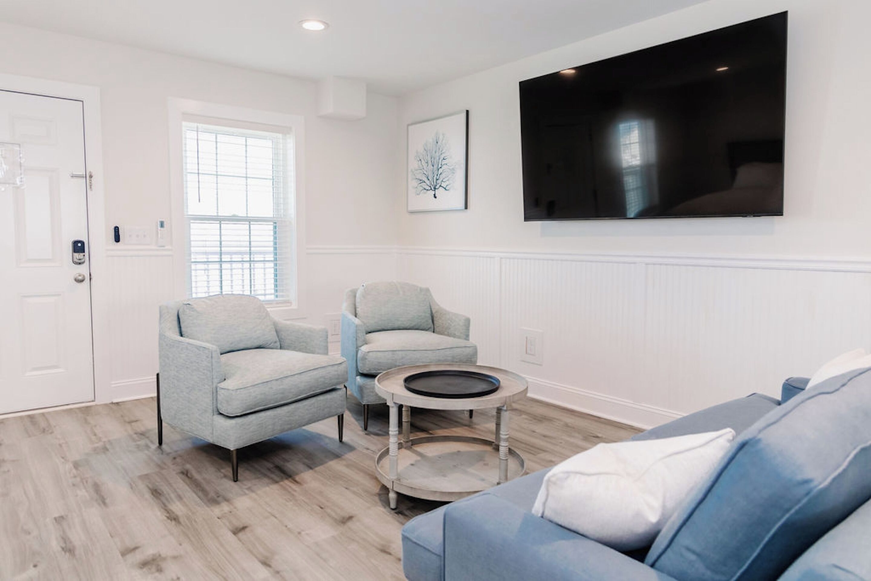 Ein Wohnzimmer mit grauem Sofa, blauem Sofa und zwei Sesseln. An der Wand ist ein Flachbildfernseher angebracht und es gibt ein Fenster mit Jalousien.