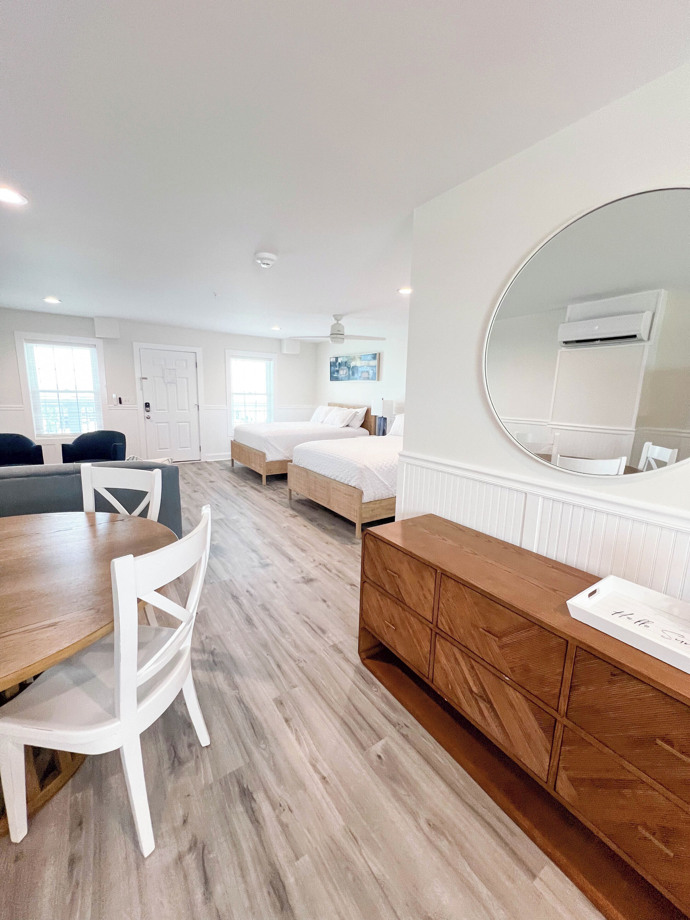 A modern hotel room with a wooden floor, a round mirror, a wooden dresser, a bed, a desk, and a chair.