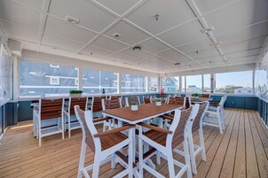 Un área de comedor al aire libre techada, con mesas de madera y sillas blancas, ventanas amplias y vistas a edificios y un barco en la distancia.