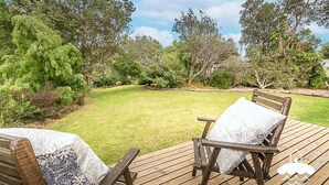 A wooden deck with two wooden chairs, each with a white cushion. The deck is surrounded by a well-maintained lawn and various trees. - Sandscape (Sandy Point)
