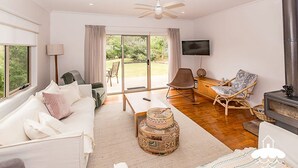 A spacious living room with a white sofa, a coffee table, and a fireplace. There is a television mounted on the wall and a large window providing a view of the outdoors. - Sandscape (Sandy Point)