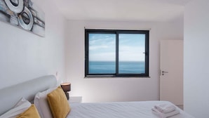 Un dormitorio con cama, mesita de noche, lámpara, ventana con vista al mar y puerta.