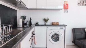Cocina compacta con armarios blancos, encimera negra, lavadora y sofá negro.