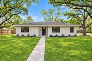 A single-story house with a front entrance, a visible number '1066' on the door, a front yard with a sidewalk, and a wooden fence in the background. - Spacious Family Gem: Private Giant Yard | 4br (Baytown)