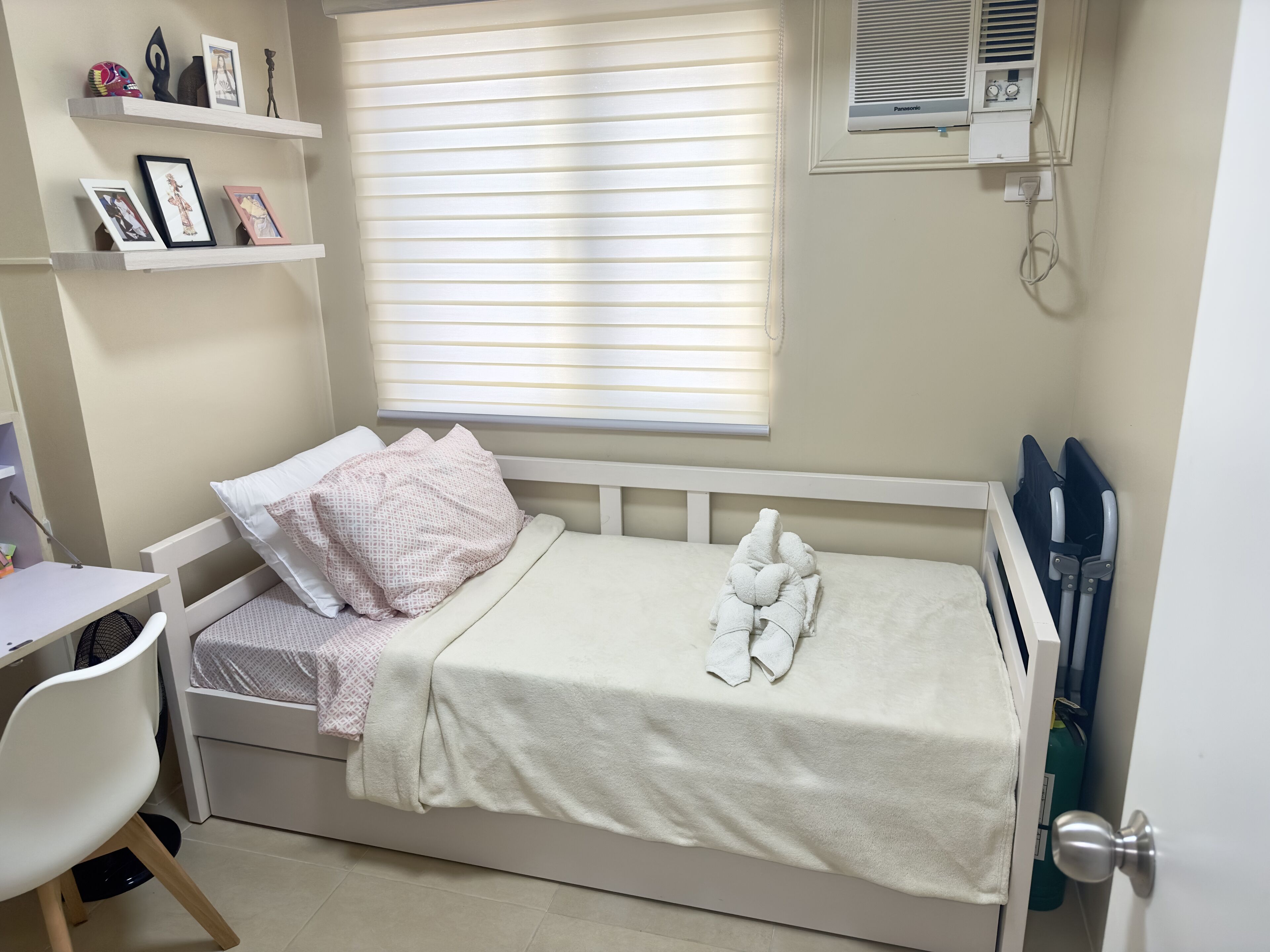 A small, neatly made bed with a white headboard, a bedside table with a chair, and a window with blinds.