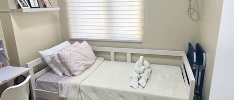 A small, neatly made bed with a white headboard, a bedside table with a chair, and a window with blinds.