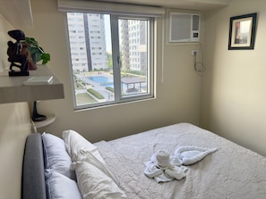 A hotel room with a bed, a window with a view of a pool and high-rise buildings, a wall-mounted air conditioner, and a phone.