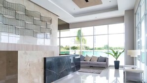A modern lobby with a marble reception desk, a grey sofa, and large windows offering a view of greenery and a pool. - Stay comfy & feel Homealike 32 (Iloilo)