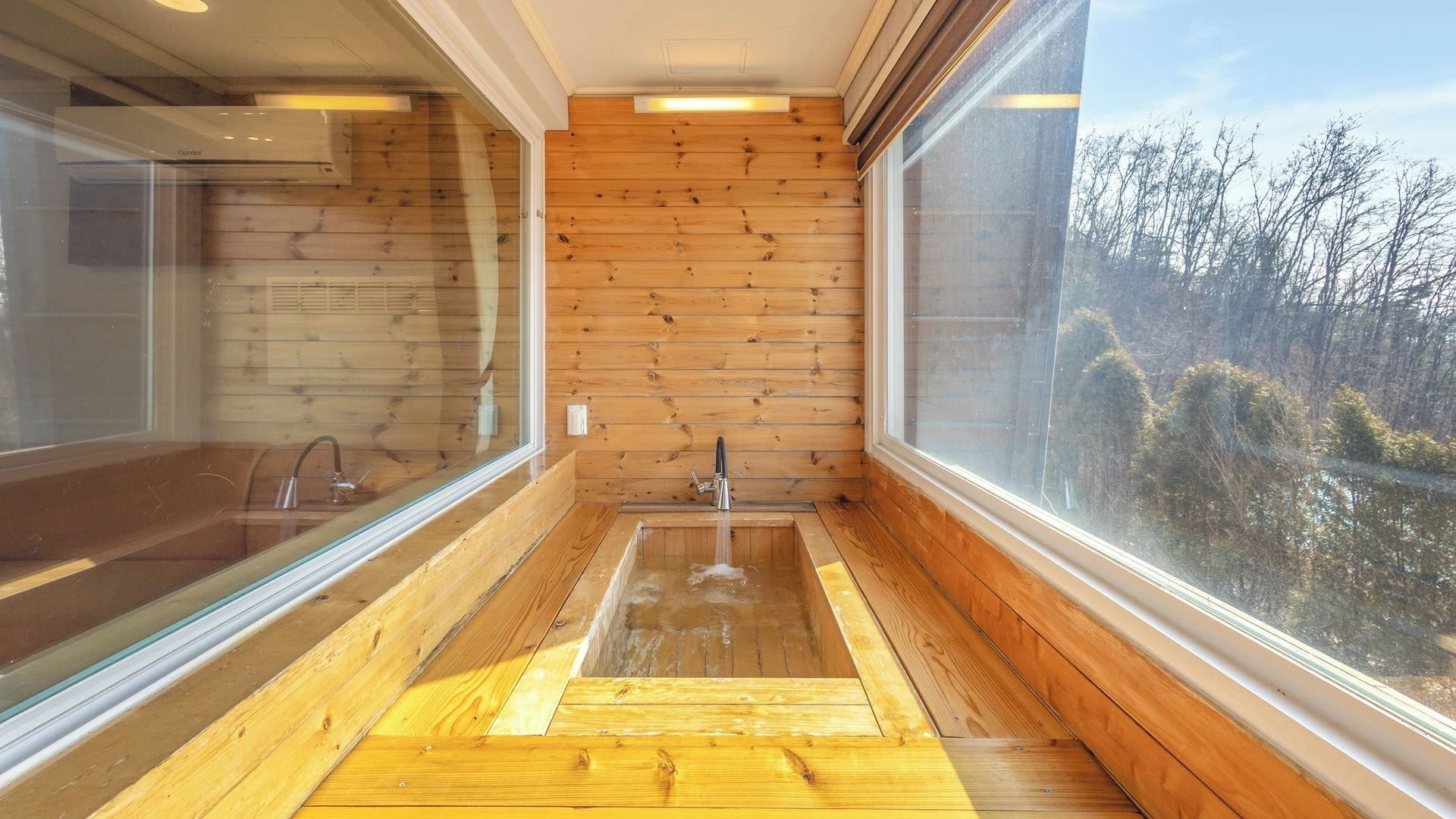 A wooden interior with a wooden floor, a sink, and large windows offering a view of trees.