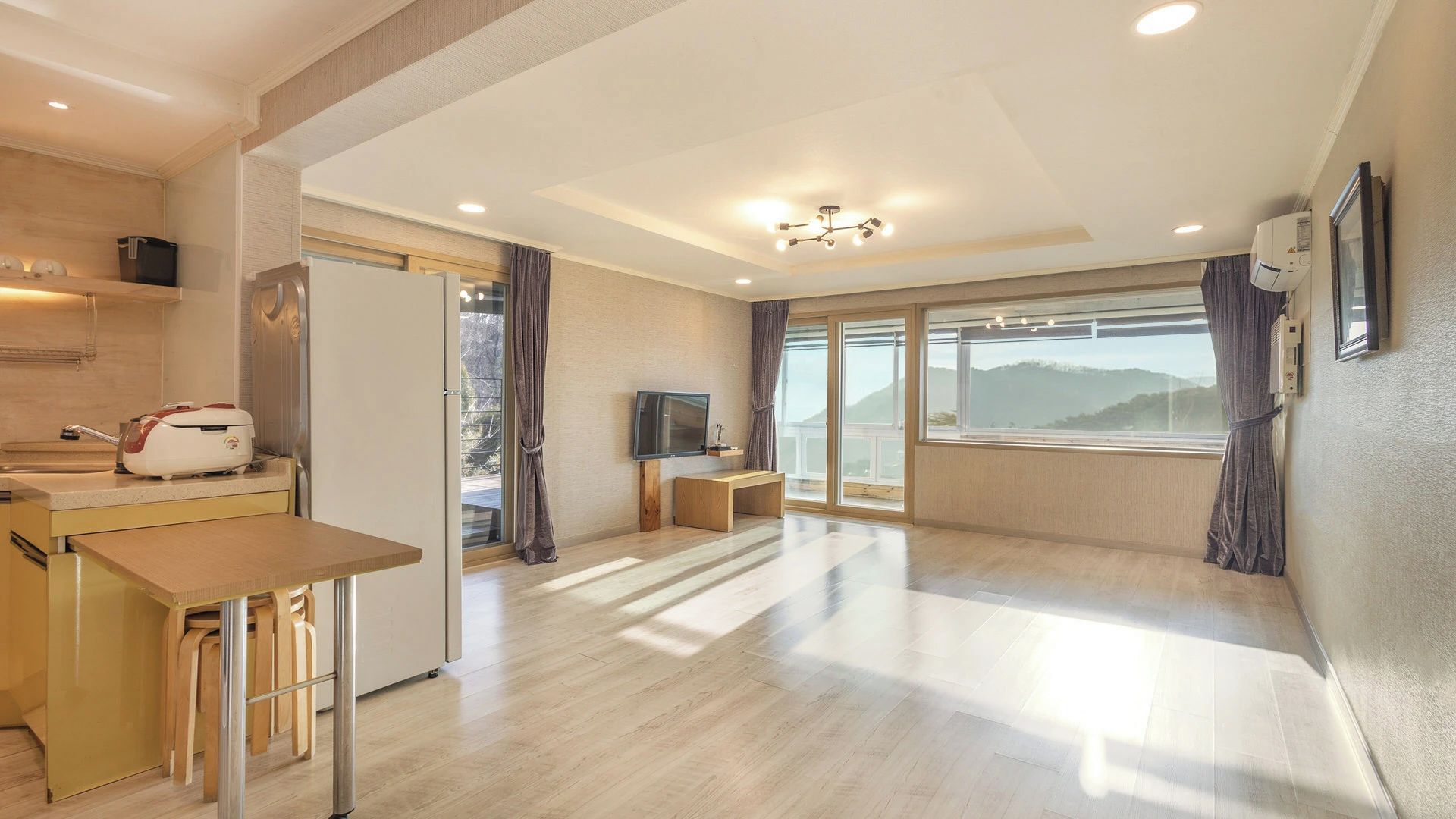 A modern hotel room with a kitchen area, a dining table, a TV, and a large window with a mountain view.