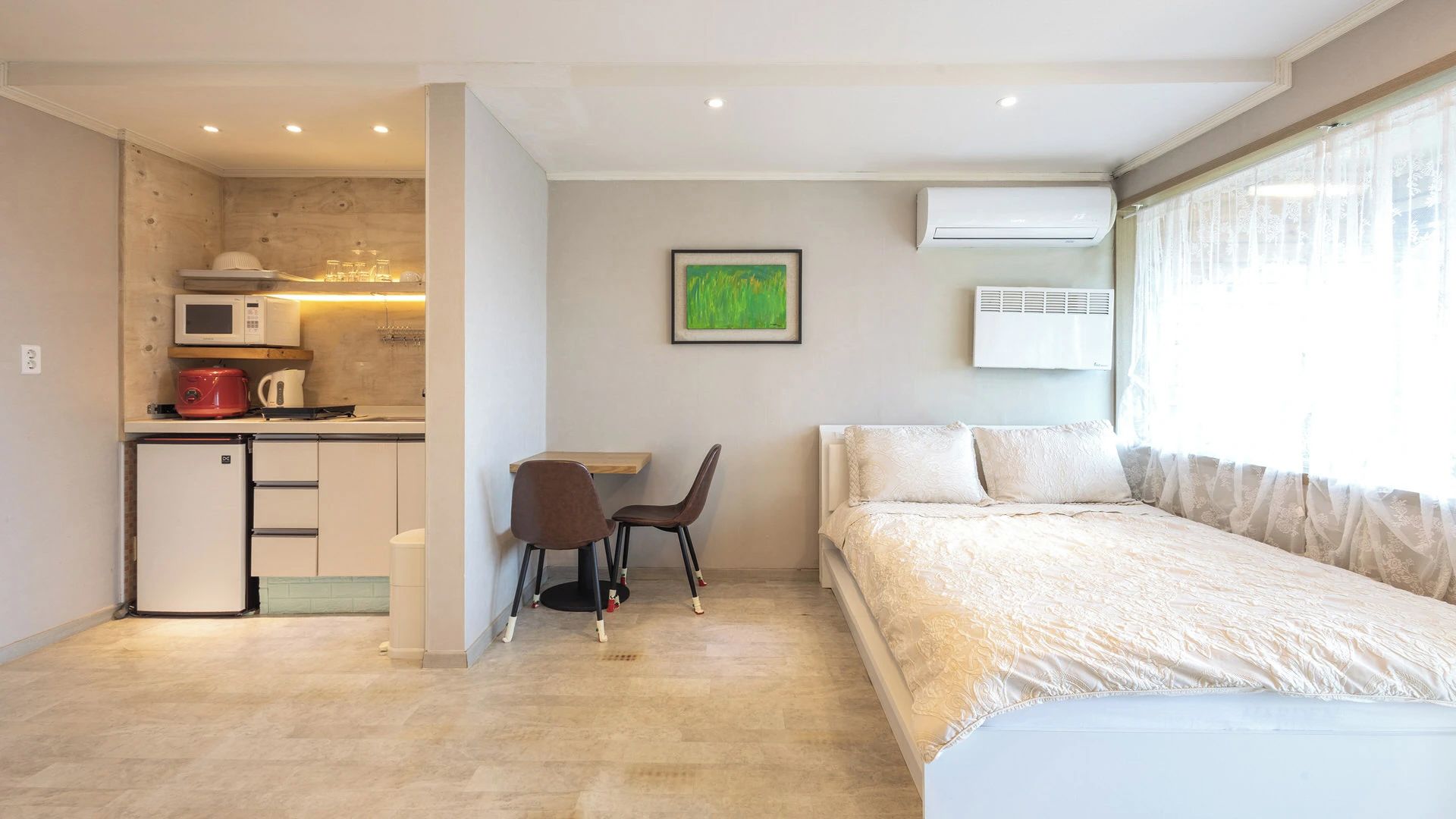 Een compacte hotelkamer met een kitchenette, een enkel bed, een bureau met stoel en een groot raam.