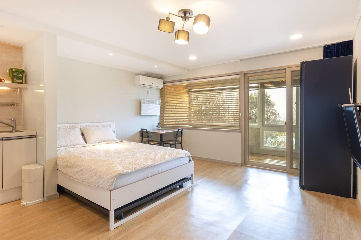 Ein modernes Schlafzimmer mit Bett, kleinem Tisch und Stuhl. Es gibt ein Fenster mit Jalousien, eine Wandklimaanlage und eine Deckenleuchte.