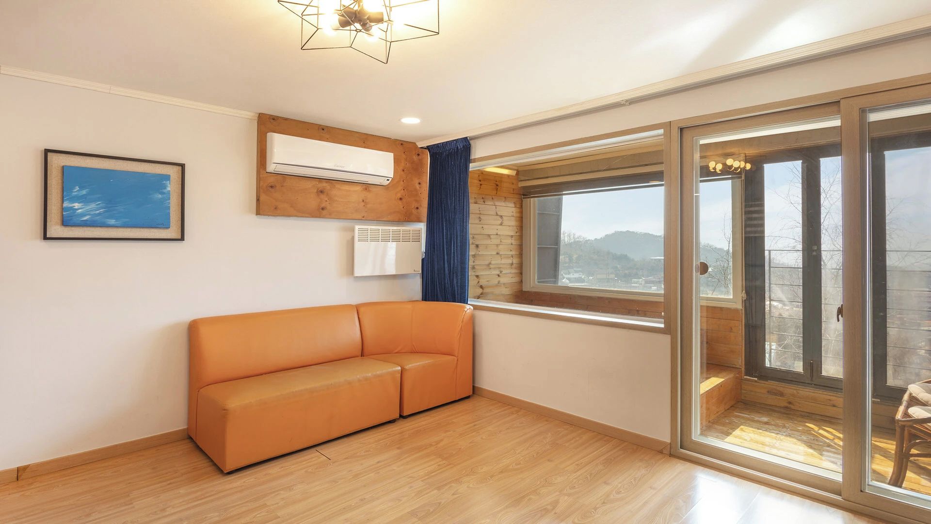 A room with a wooden floor, a sofa, a window with a view, and an air conditioning unit.