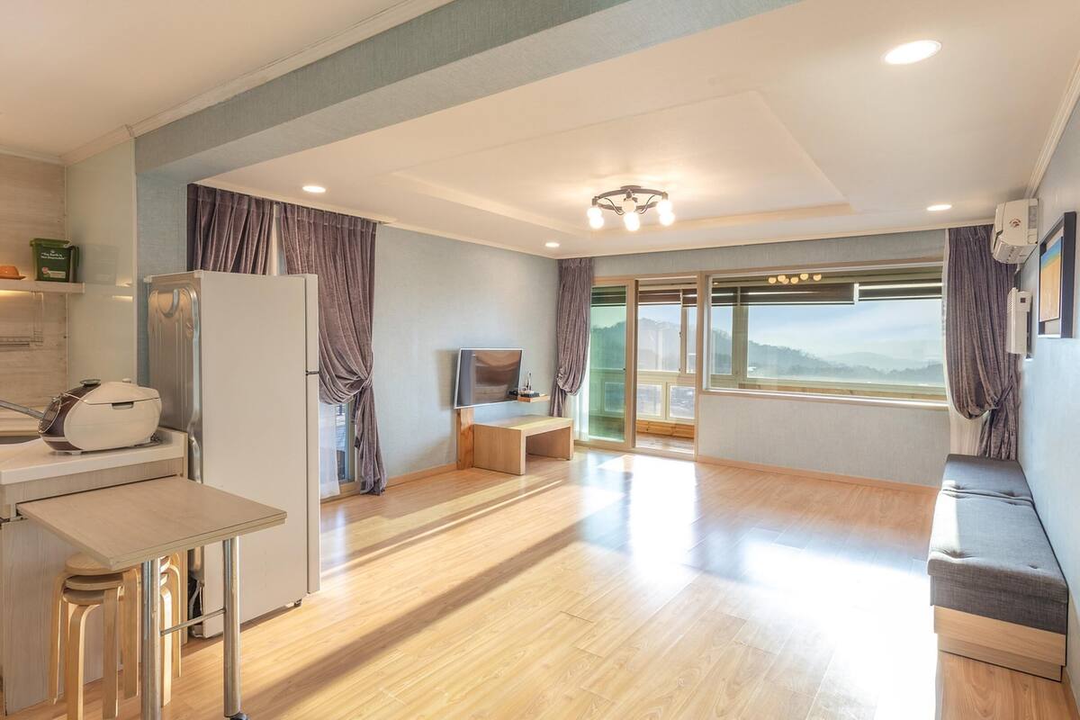 Eine moderne Küche mit Kühlschrank, Herd und Spüle. Ein Holztisch und eine Bank bieten Platz. Große Fenster mit Vorhängen ermöglichen einen Blick ins Freie.