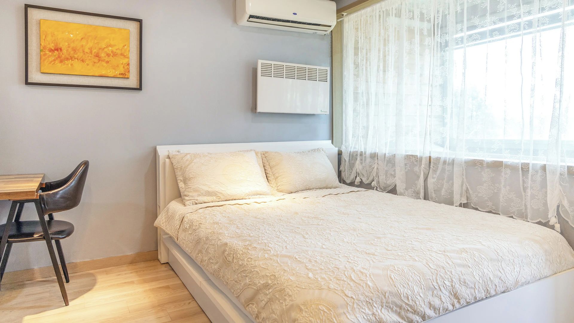 Une chambre à coucher comprenant un lit, une chaise, un appareil de climatisation et une fenêtre avec des rideaux légers.