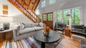 A spacious living room with a high ceiling, wooden staircase, large windows, and a cozy seating area. - Modern Log Cabin - Hot Tub, Fire Pit, Spa-like bathroom in secluded nature haven (Blue Ridge)