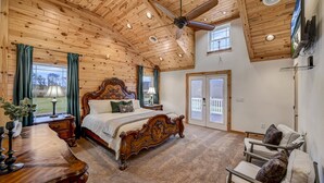 Un dormitorio con techo de madera, una cama grande, ventilador de techo, televisor y vistas al exterior.