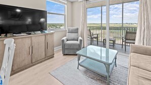 Ein modernes Wohnzimmer mit Flachbildfernseher, grauem Sessel, Glastisch und einem Balkon mit Gartenmöbeln.