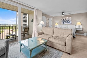 Habitación de hotel con balcón, sofá, mesa de centro de vidrio, cama y dos lámparas de noche.