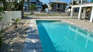 A rectangular swimming pool with clear blue water, surrounded by a white fence and a gravel area.