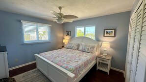 Une chambre à coucher comprenant un lit, deux tables de chevet, un ventilateur de plafond et deux fenêtres équipées de stores.