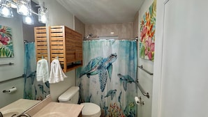 Une salle de bain avec un rideau de douche orné de motifs de tortues de mer, une étagère en bois au-dessus des toilettes et un miroir.