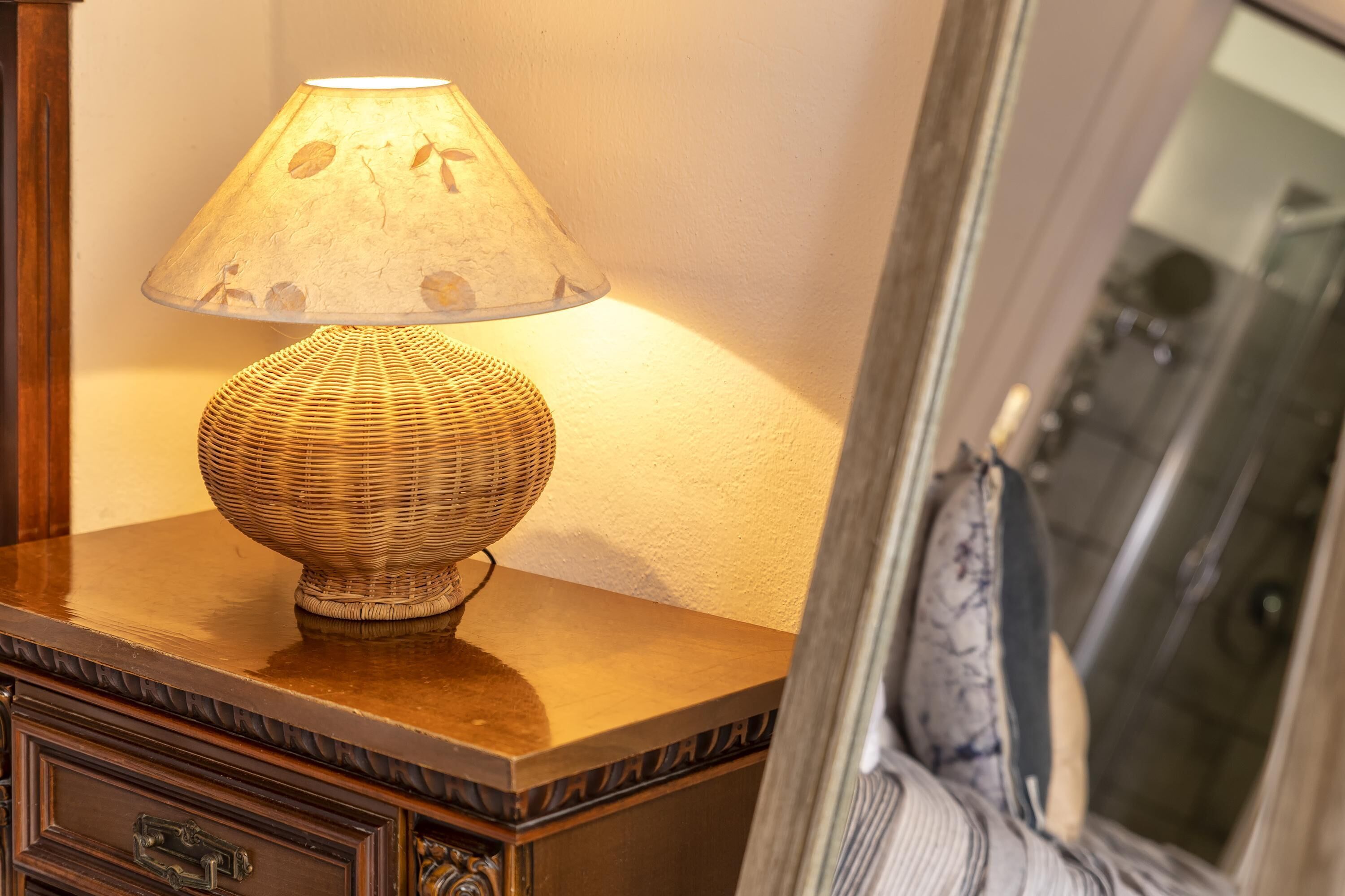 Une lampe dont le pied est fait d’un matériau tissé et l’abat-jour est orné d’un motif de feuilles, posée sur une table en bois.