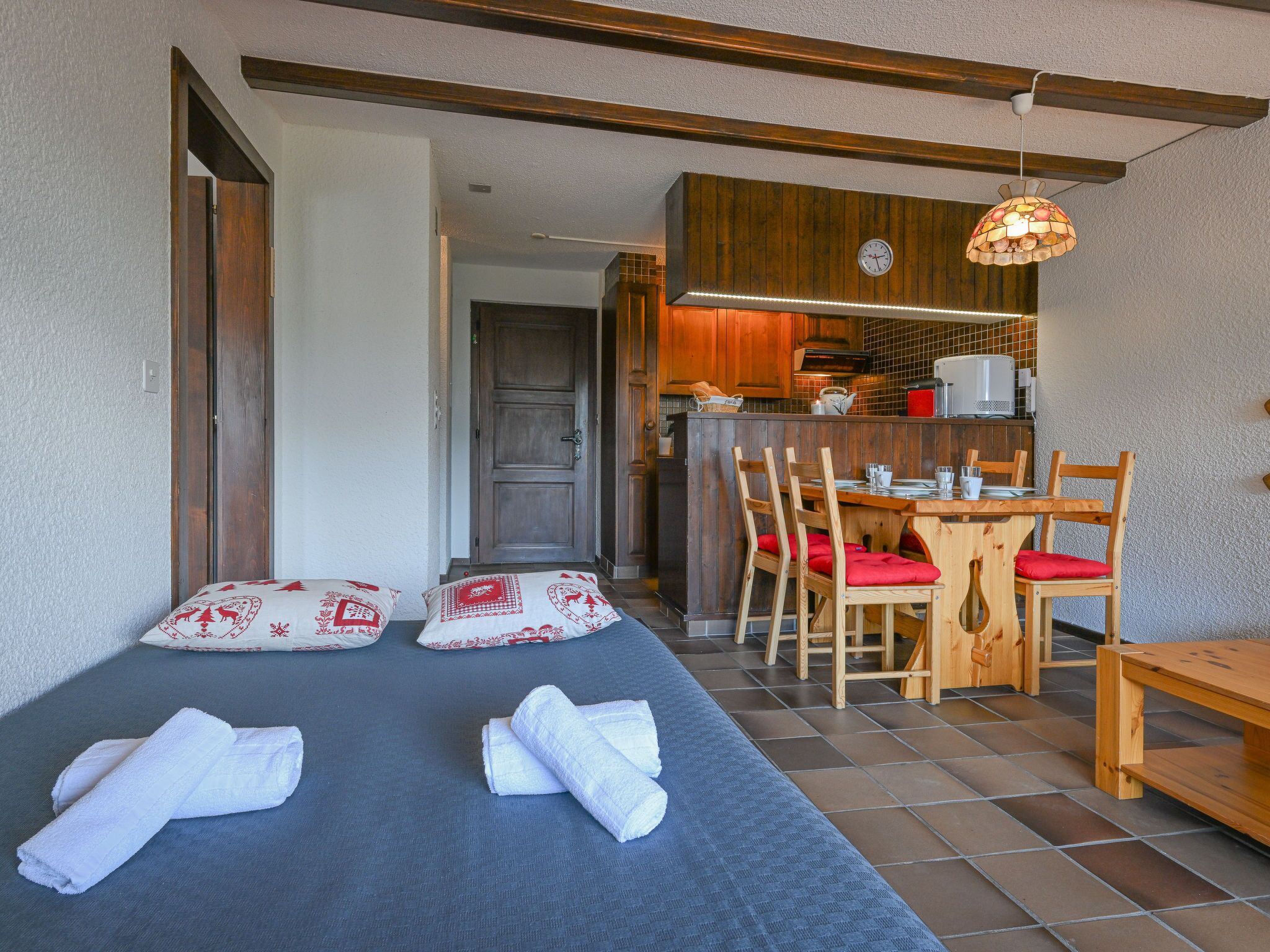 Une pièce comprenant un lit, une table à manger en bois avec des chaises, un coin cuisine et une horloge au mur.