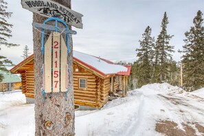 Een houten chalet met een rood dak, omgeven door sneeuw en bomen. Een bord met de cijfers 211 en 57 zit vast aan een boom.