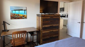 Ein Schlafzimmer mit Bett, Schreibtisch mit Stuhl, Kommode, Fernseher und einem durchgehenden Durchgang erkennbaren Küchenbereich.