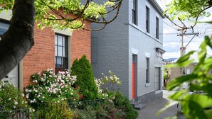 En boligbygning med facade af mursten, en grå overdel og en rød dør. Der er en have med forskellige planter og blomster foran.