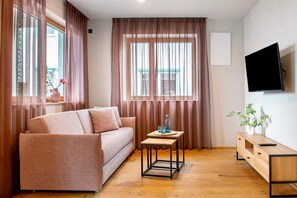 Una sala de estar con sofá, una mesita de madera, un televisor montado en la pared y un ventanal con cortinas.