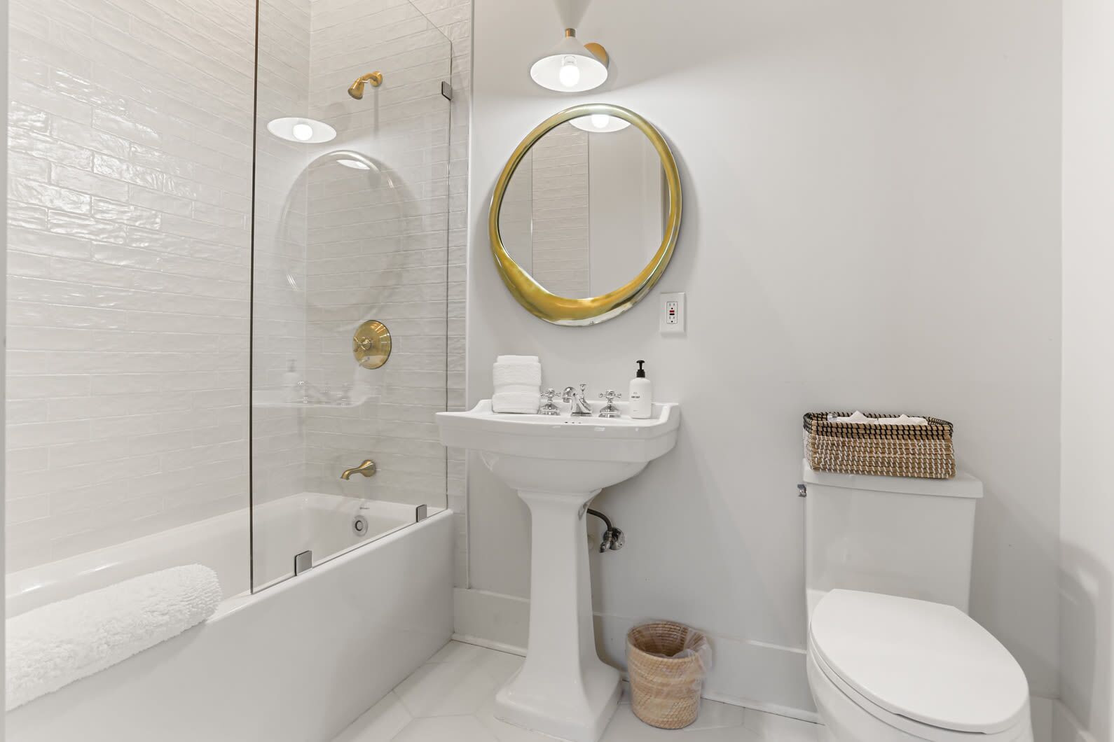 Een moderne badkamer met een douche met glazen wand, een wastafel op een zuil, een toilet en een rieten mand.