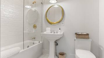 Een moderne badkamer met een douche met glazen wand, een wastafel op een zuil, een toilet en een rieten mand.