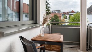 Ein Balkon mit einem Holztisch und Stuhl, einer Vase mit getrockneten Pflanzen und Blick auf Gebäude und Bäume.