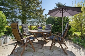 Un espace extérieur aménagé avec des meubles en bois, comprenant une table, des chaises et un parasol, le tout agrémenté d’un jardin en arrière-plan.