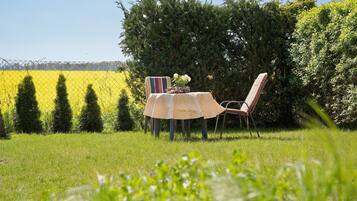A table with a tablecloth and chairs in a grassy area with a hedge and trees in the background.