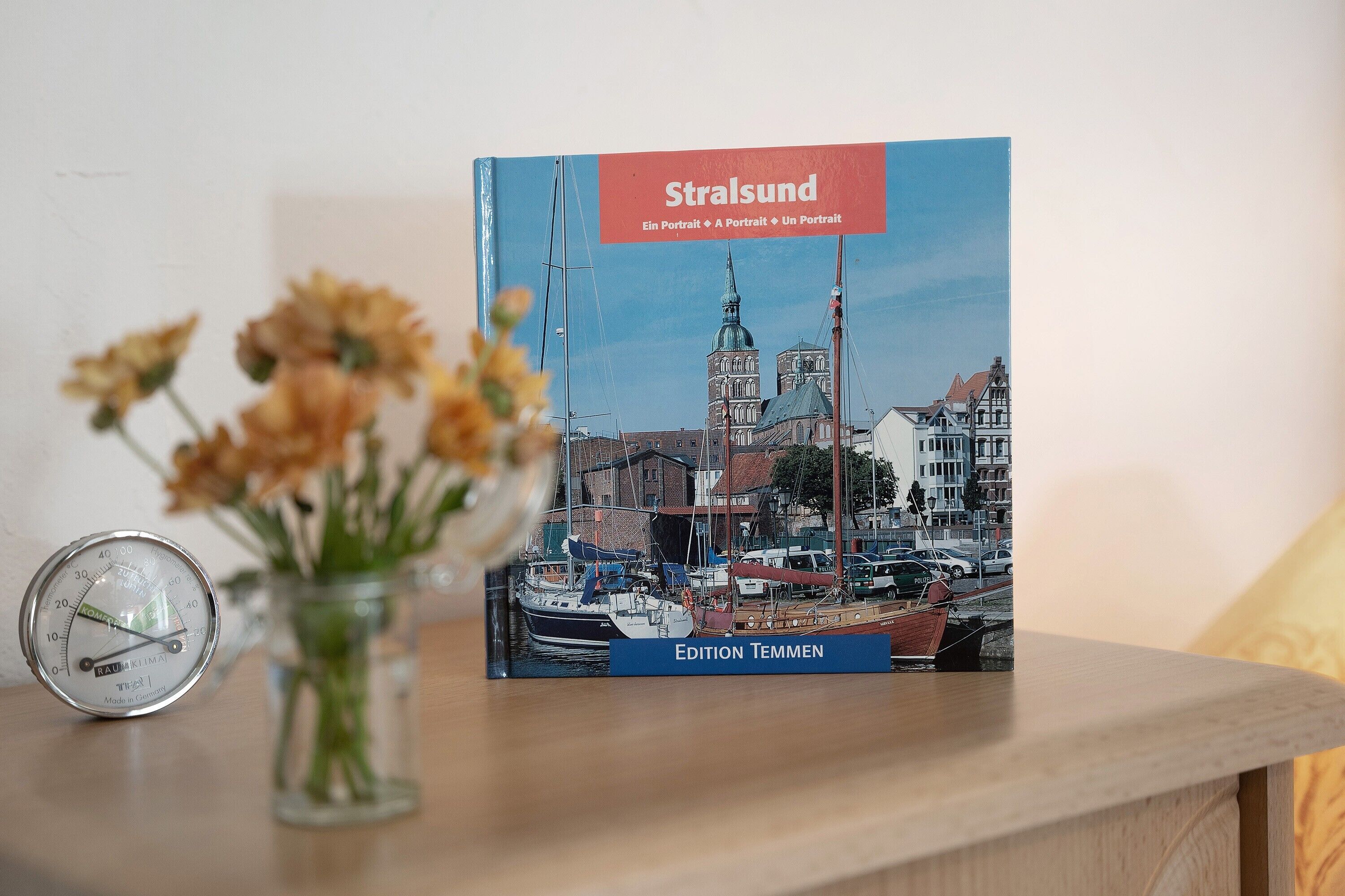 Puupöydällä on kirja, jonka nimi on ”Stralsund”. Kirjan vieressä on lasivaasi, jossa on kukkia, sekä lämpömittari.