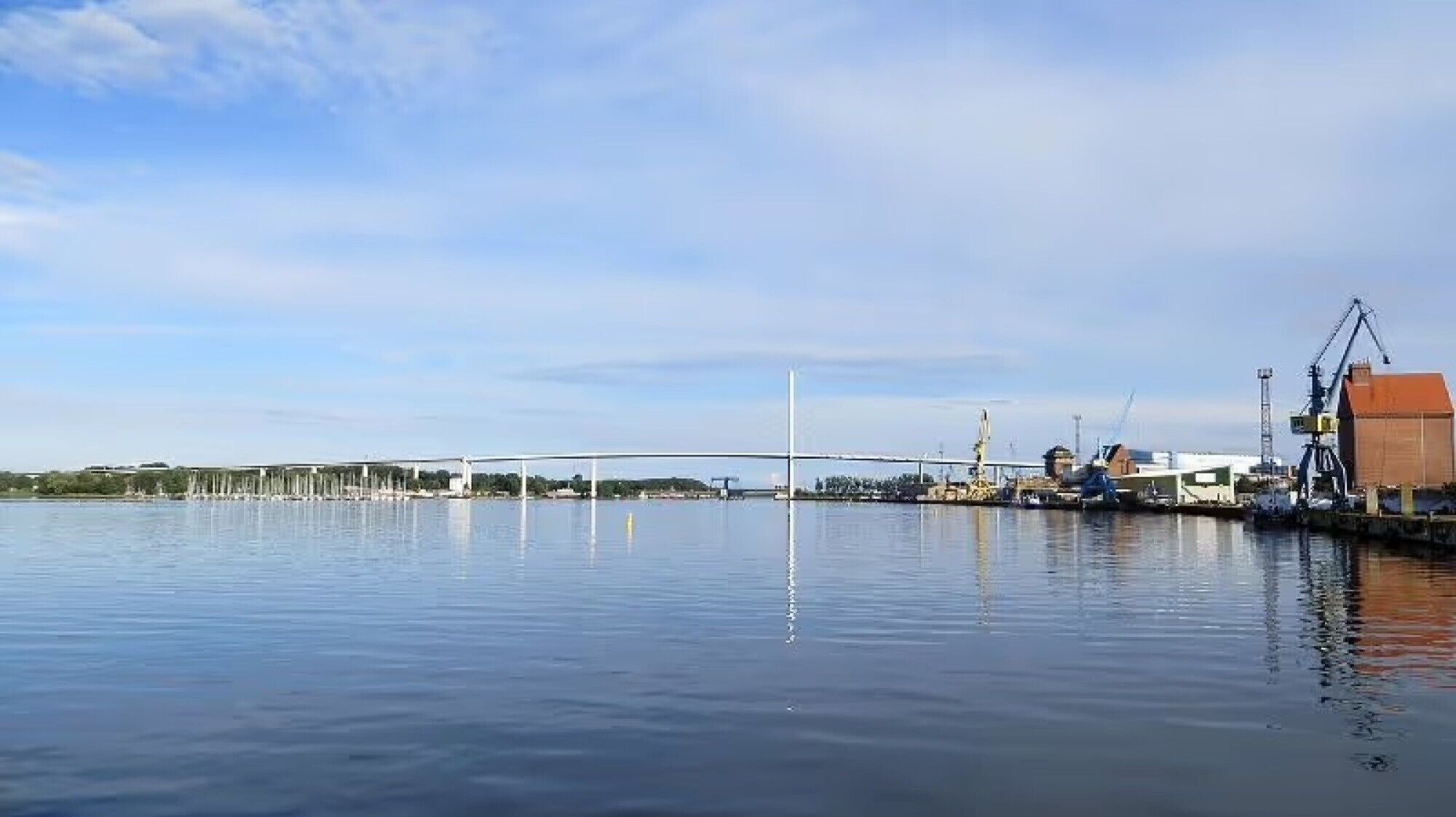 A calm waterfront with a bridge, cranes, and industrial buildings.