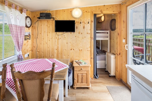 Un intérieur en bois comprenant une table à manger, un téléviseur, un petit meuble et un lit superposé, visibles à travers une porte ouverte.