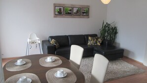 Ein Wohnzimmer mit einer schwarzen Couch, einem Esstisch mit Platzsets, einem weißen Stuhl, einem kleinen weißen Tisch und einer Pflanze.