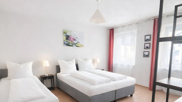 Ein Hotelzimmer mit zwei Betten, Holzboden, Deckenleuchte, einem Fenster mit Vorhängen und einem Wandbild.
