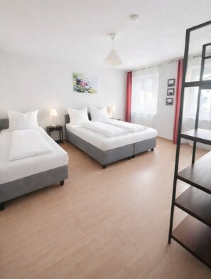 Ein Hotelzimmer mit zwei Betten, Holzboden, Deckenleuchte, einem Fenster mit Vorhängen und einem Wandbild.