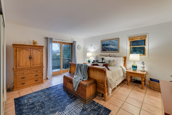 A bedroom with a bed, nightstands, a wooden dresser, a window with a view, and a door leading to a balcony.